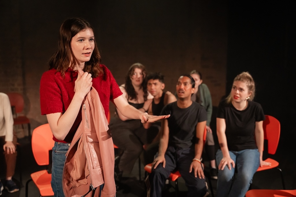 Action shot of the spring 2024 Shakespeare in Performance at RADA final presentation of Julius Caesar. An actor wearing a bright red shirt holds a light pink jacket in mid-performance as other actors, seated, look on. 